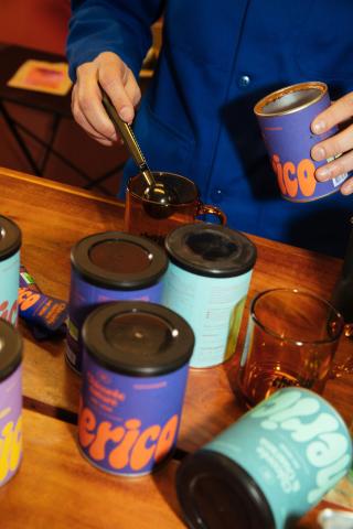 Person scooping instant coffee powder into a glass cup with colorful Cherico tins on a wooden counter