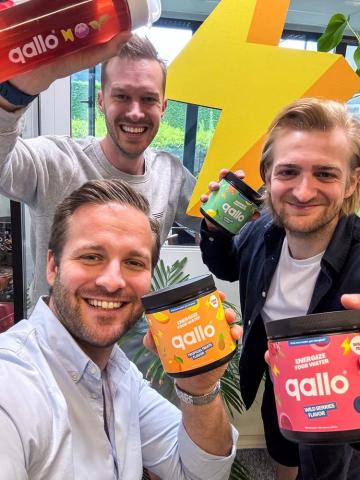 Three men smiling and holding Qallo drink mix containers in an office environment