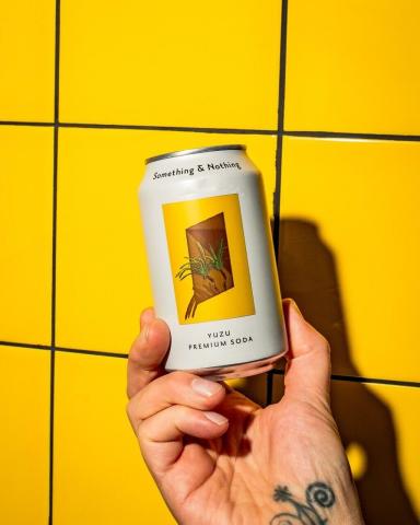 Hand holding a Something & Nothing yuzu premium soda can against a yellow tiled background